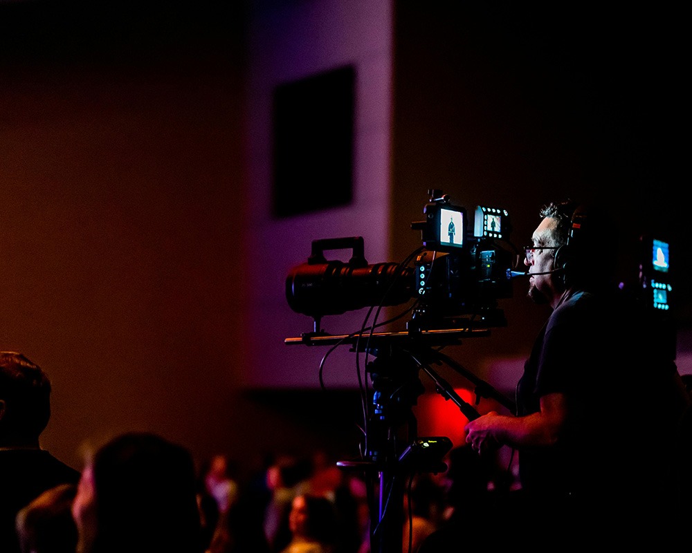 A professional camera operator wearing a headset operates a high-definition broadcast video camera on a tripod, filming a live corporate event or hybrid conference from the back of a dimly lit venue.