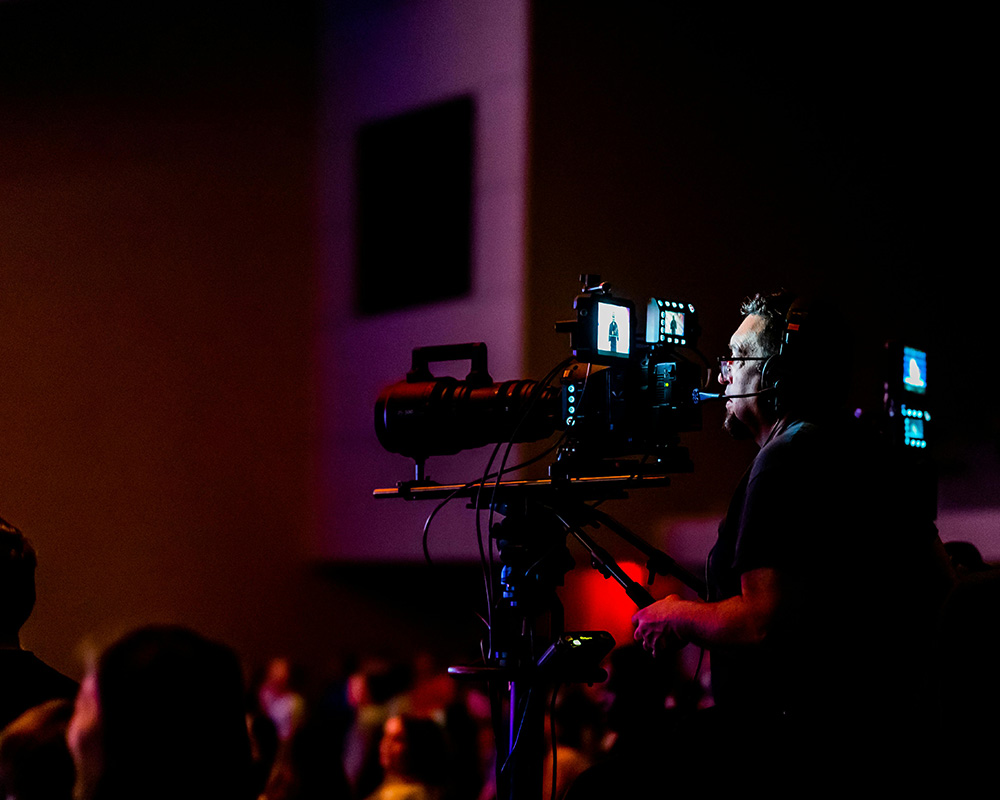 A professional camera operator wearing a headset operates a high-definition broadcast video camera on a tripod, filming a live corporate event or hybrid conference from the back of a dimly lit venue.